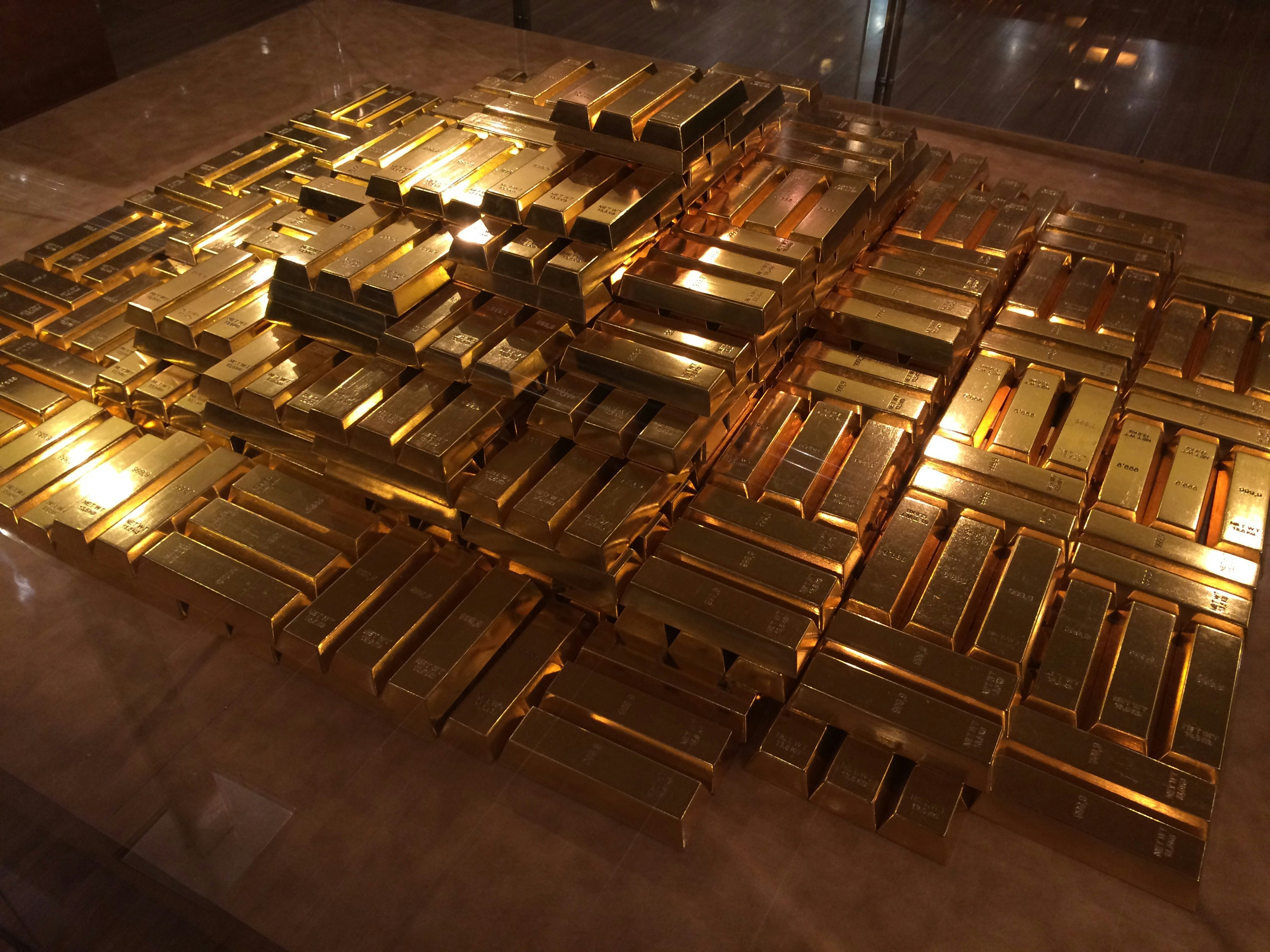 A large stack of shiny gold bars neatly arranged on a reflective surface, creating an impression of wealth and opulence. The bars are aligned in rows, with light glinting off their polished surfaces.