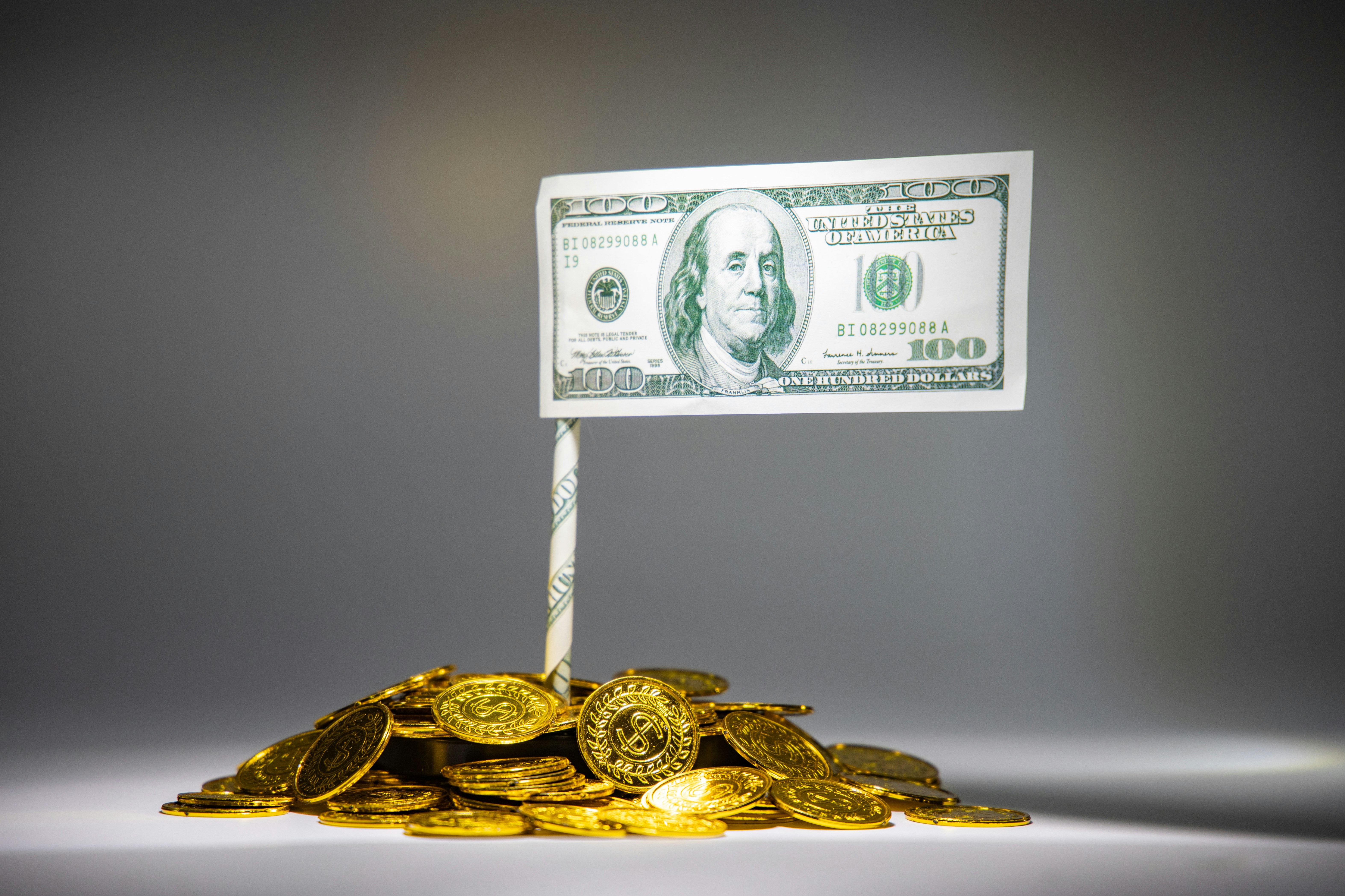 A meticulously arranged pile of shiny gold coins is topped by a rolled paper acting as a pole that holds a one hundred dollar bill. The setup resembles a flag mounted on a mound of wealth.